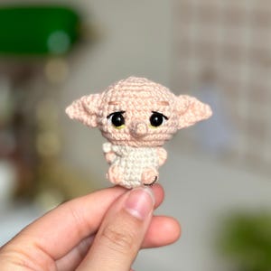 Può includere: Un piccolo giocattolo amigurumi all'uncinetto rosa di un elfo domestico con occhi grandi e orecchie a punta. L'elfo tiene le mani davanti a sé.