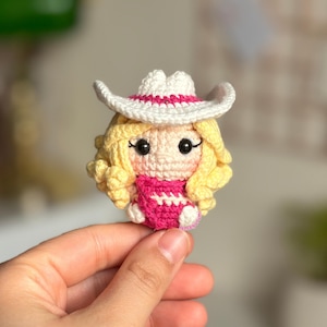 Könnte beinhalten: Eine gehäkelte Puppe mit einem weißen Cowboyhut mit rosa Band, einem rosa-weiß gestreiften Shirt und gelben Locken.