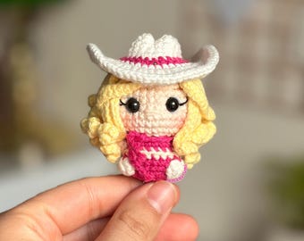 Peut inclure: Une poupée en crochet portant un chapeau de cow-boy blanc avec un bandeau rose, une chemise rayée rose et blanche et des cheveux bouclés jaunes.