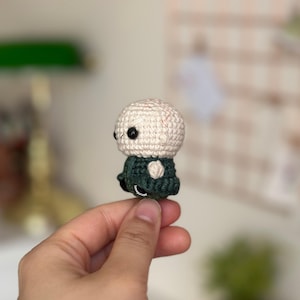 May include: A small crocheted figure of a bald person wearing a green jacket. The figure is being held in a hand.
