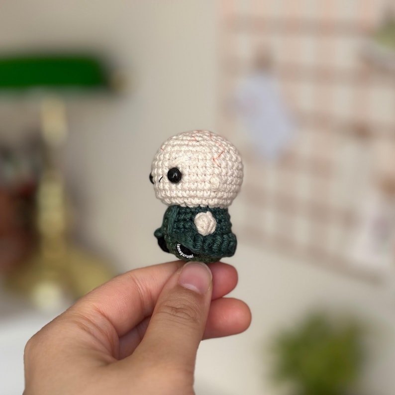 May include: A small crocheted figure of a bald person wearing a green jacket. The figure is being held in a hand.