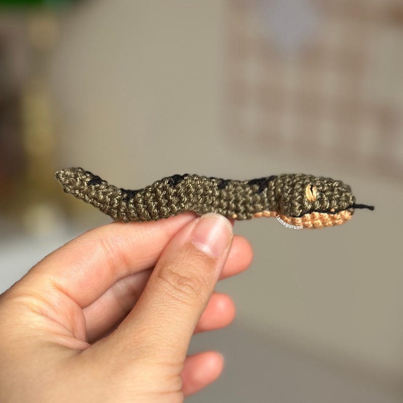 May include: A small crocheted snake toy with a green and black body and a light brown head. The snake is being held in a person's hand.