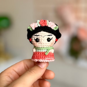 Puede incluir: Una muñeca de crochet con cabello negro, un vestido rosa y verde y una corona de flores. La muñeca tiene una cara pintada con ojos marrones y un ligero ceño fruncido.