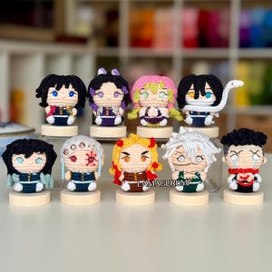 May include: A collection of nine small, handcrafted crochet dolls featuring various anime-inspired characters with distinct hair colors, facial expressions, and outfits. Each figure is mounted on a small, round wooden base. The text LAMAGURUMI is visible.