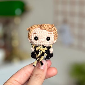 Peut inclure: Une poupée en crochet amigurumi représentant un jeune garçon aux cheveux bruns, portant une écharpe noire et jaune. La poupée est tenue dans une main.
