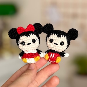 May include: Two crocheted amigurumi dolls, one with a red bow and the other with red shorts, both with black ears and white faces.