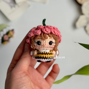 Könnte beinhalten: Eine handgefertigte Häkelpuppe mit einem rosa Blumenhut, braunen Haaren und einem gelb-schwarz gestreiften Kleid. Die Puppe hat große schwarze Augen und hält einen kleinen grünen Stiel. Das Wort "LAMAGURUMI" ist auf dem Kleid sichtbar.