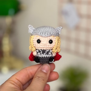 Op de afbeelding: Een handgemaakte Thor-pop gehaakt. De pop heeft een grijze helm, blond haar en een rode cape. De outfit is zwart en grijs. De pop is klein en lijkt een verzamelobject te zijn.