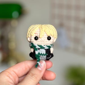 Puede incluir: Una muñeca amigurumi de crochet de una persona joven con cabello rubio, que lleva una bufanda verde y blanca y un atuendo negro.