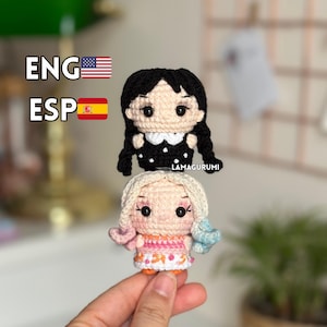 Peut inclure: Deux poupées au crochet faites à la main. La poupée du haut a des cheveux noirs tressés, un col blanc et une robe noire à pois blancs. La poupée du bas a des cheveux blonds avec des accents roses et bleus et une robe orange et rose. Le texte "LAMAGURUMI" est visible.