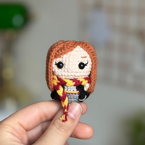 Könnte beinhalten: Eine gehäkelte Amigurumi-Puppe einer jungen Frau mit roten Haaren, die einen rot-goldenen Schal trägt. Die Puppe hält einen Zauberstab in der Hand.