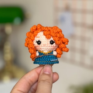 以下が含まれることがあります： オレンジ色の髪、青いドレス、茶色の目をした、かぎ針編みの人形。人形は、茶色のロープを手に持っています。人形の高さは約7.5cmです。