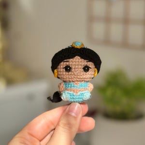 Puede incluir: Muñeca de ganchillo hecha a mano de una princesa con cabello negro, un atuendo azul y una tiara dorada. La muñeca tiene grandes ojos negros y pequeños pendientes dorados. La muñeca se sostiene en una mano.