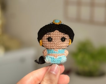 May include: A handmade crochet doll of a princess with black hair, a blue outfit, and a gold tiara. The doll has large black eyes and small gold earrings. The doll is held in a hand.