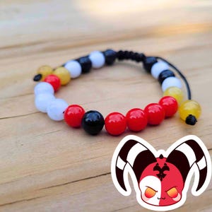 May include: A beaded bracelet featuring a mix of red, black, white, and yellow beads. The bracelet is strung on a black cord and rests on a wooden surface. A cartoon image of a red and white character is in the lower right corner.