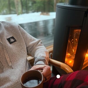 Könnte beinhalten: Eine Person sitzt auf einem Stuhl vor einem Kamin und trägt ein graues Hoodie mit einem schwarzen Patch und eine rot-schwarze karierte Hose. Sie hält eine braune Tasse mit einer dunklen Flüssigkeit.