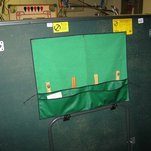 May include: A green felt-covered table with a green fabric pocket on the side. The pocket has four wooden dowels sticking out of it. The table has a black metal frame and a black metal leg.