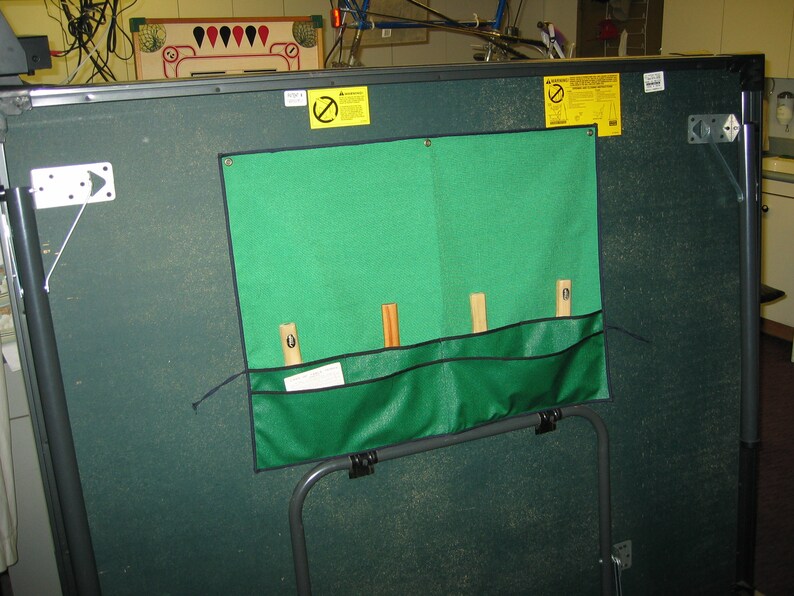 May include: A green felt-covered table with a green fabric pocket on the side. The pocket has four wooden dowels sticking out of it. The table has a black metal frame and a black metal leg.