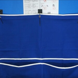 May include: A blue canvas storage bag with white trim and three pockets hangs on a table tennis table. The bag has two black straps with metal eyelets.