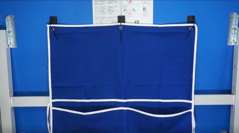 May include: A blue canvas storage bag with white trim and three pockets hangs on a table tennis table. The bag has two black straps with metal eyelets.
