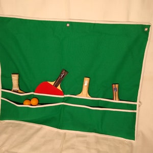 May include: A green canvas wall organizer with white trim and four pockets. The organizer has three wooden table tennis paddles and two orange table tennis balls in the top pocket.