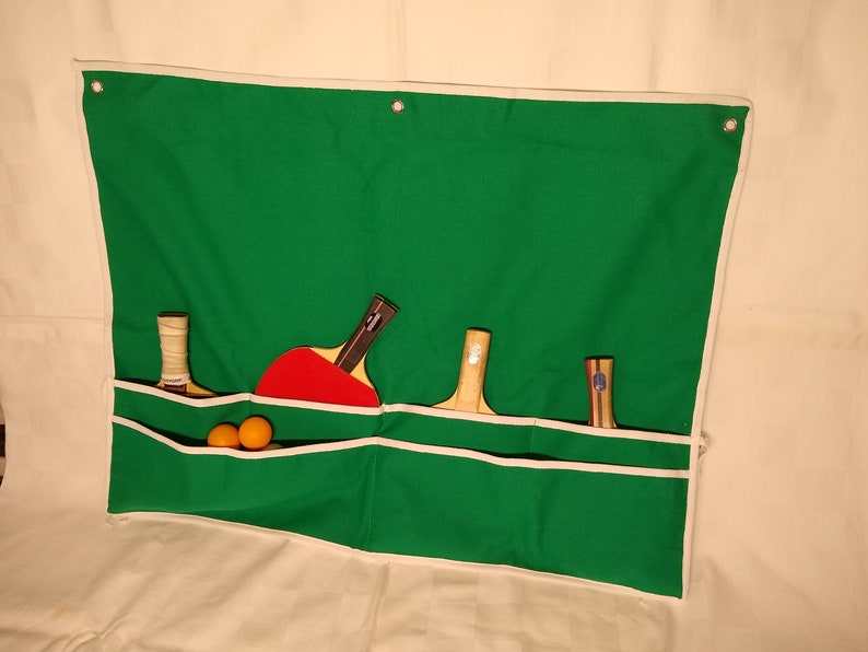 May include: A green canvas wall organizer with white trim and four pockets. The organizer has three wooden table tennis paddles and two orange table tennis balls in the top pocket.