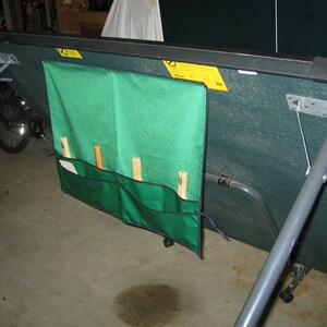 May include: A green and gray folding table tennis table with a green storage bag attached to the side. The bag has four pockets for storing paddles and balls. The table is folded up and resting on its legs.