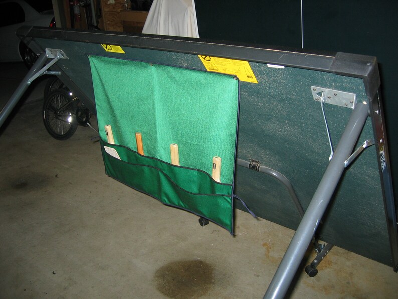 May include: A green and gray folding table tennis table with a green storage bag attached to the side. The bag has four pockets for storing paddles and balls. The table is folded up and resting on its legs.