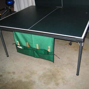 May include: A black and green folding table tennis table with a green storage bag attached to the side. The bag has three compartments for storing paddles and balls.
