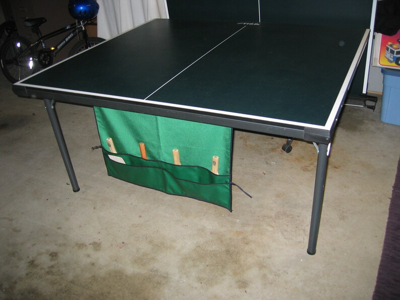 May include: A black and green folding table tennis table with a green storage bag attached to the side. The bag has three compartments for storing paddles and balls.