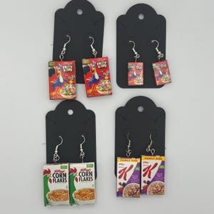 May include: A collection of novelty earrings featuring miniature cereal boxes. The earrings include designs from Kellogg's Froot Loops, Corn Flakes, and Special K cereal boxes. Each pair is attached to a black earring display card.