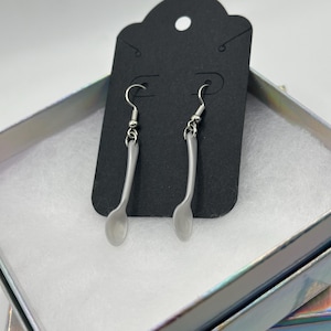 May include: A pair of silver-colored spoon earrings displayed on a black earring card. The earrings are made of a shiny metal and have a simple, minimalist design. The earrings are presented in a decorative box with a white interior.