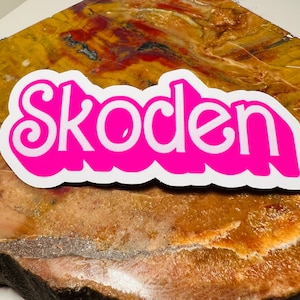 Könnte beinhalten: Ein rosa Aufkleber mit dem Wort "Skoden" in weißen Buchstaben. Der Aufkleber befindet sich auf einer braunen und gelben gemusterten Oberfläche.