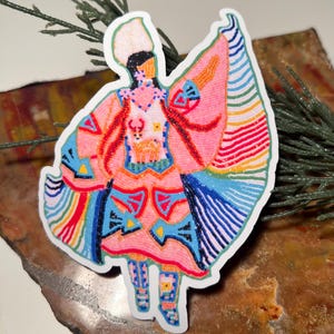Op de afbeelding: Een kleurrijke sticker met een kralenversierde indiaanse danseres die een roze jurk draagt met geometrische patronen en een witte hoofdtooi. De danseres wordt omringd door een regenboog van kleuren.