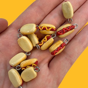 May include: A collection of miniature hot dog charms. The charms feature a bun, a red hot dog, and yellow mustard. Some charms are just the bun. Each charm has a silver-colored loop for attaching to jewelry or other crafts.