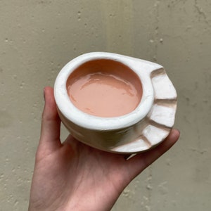May include: A white ceramic planter with a ruffled edge and a light pink interior. The planter is designed to resemble a seashell.