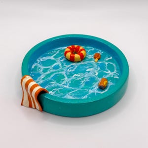 May include: A miniature pool scene with a turquoise rim and water effect. Inside are a yellow and red inflatable ring, a popsicle, a can, and a towel with orange and white stripes. A fun, decorative piece.