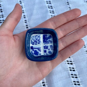 Op de afbeelding: Een kleine, vierkante, blauwe keramische schaal met vier witte vierkanten. Elk wit vierkant heeft een ander blauw bloemen- of abstract patroon. De schaal wordt in een hand gehouden.