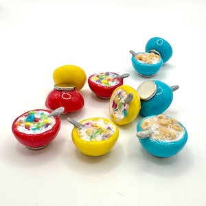 May include: A collection of miniature bowl-shaped magnets featuring colorful cereal in milk with a small spoon. The bowls are painted in bright red, yellow, and turquoise blue. Each piece includes a metallic circular magnet on the back side.