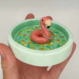May include: A round, light mint green ceramic trinket dish featuring a pool-themed interior with a grid of blue, green, and yellow tiles. A pink flamingo figure sits in the center, holding a thin silver ring with small embedded stones.