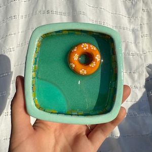 Pode incluir: Um prato quadrado de cerâmica com interior turquesa e borda verde claro. Um flutuador de anel de cerâmica laranja, decorado com flores, repousa dentro do prato. O prato é segurado em uma mão.