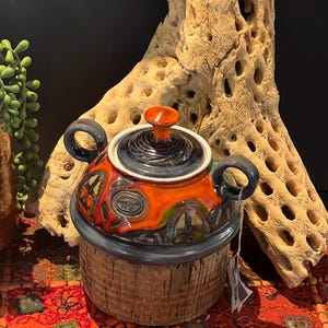 May include: A ceramic jar with a lid, featuring a colourful, abstract design in shades of orange, blue, and green. The jar has two handles and is sitting on a red and orange patterned rug.
