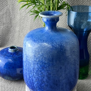 Könnte beinhalten: Eine blaue Keramikvase mit texturierter Glasur. Die Vase hat einen breiten, runden Körper und einen schmalen Hals. Sie steht auf einem weißen Sockel.