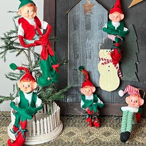 Könnte beinhalten: Fünf vintage Weihnachts-Elfen-Ornamente mit grüner und roter Filzkleidung. Die Elfen sitzen auf einem kleinen Weihnachtsbaum, einem Schneemann und einem Holzhaus. Die Ornamente befinden sich auf einem gemusterten Teppich.