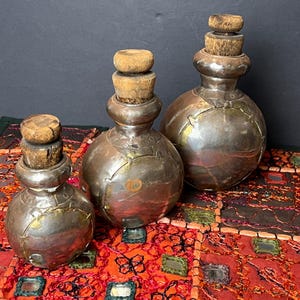 Puede incluir: Tres botellas de metal plateado con tapones de madera. Las botellas son redondas y tienen una superficie texturizada. Están sobre una tela colorida con un patrón geométrico.