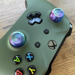 Può includere: Primo piano di un controller Xbox verde con levette colorate e vorticose. Il controller ha pulsanti neri con simboli e lettere colorate. Il logo Xbox è visibile.
