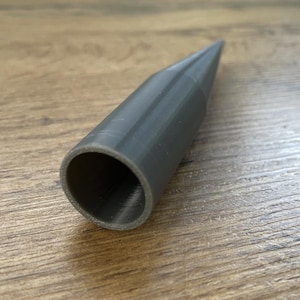 Könnte beinhalten: Ein dunkelgraues, zylindrisches Objekt mit konischer Spitze. Das Objekt hat einen hohlen Innenraum und scheint aus einem festen Material zu bestehen. Das Objekt liegt auf einer Holzoberfläche.