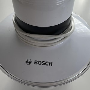 Puede incluir: Base de electrodoméstico blanca y negra con un cable blanco enrollado. La base tiene un borde plateado y el logotipo de Bosch impreso en negro. El electrodoméstico es probablemente un electrodoméstico de cocina o para el hogar.
