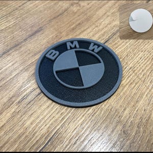 Op de afbeelding: Een zwart en grijs BMW-logo-embleem. Het cirkelvormige embleem heeft de letters "BMW" in grijs, een grijs en zwart geblokt centrum en een grijze rand. Een witte sticker bevindt zich in de rechterbovenhoek.