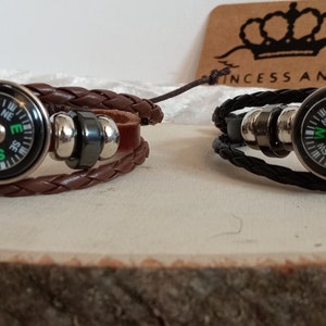 May include: Two leather bracelets with compass charms. The bracelet on the left is brown leather with a silver compass charm with a green needle. The bracelet on the right is black leather with a silver compass charm with a green needle. The bracelets are on a wooden surface.